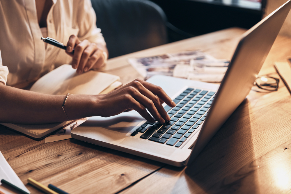 Simple and easy consultant accounting Person typing on a laptop at a wooden desk with a notebook, pen, and scattered photos nearby